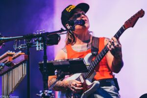Female guitarist performing live on stage, singing into a microphone with an orange tank top and a cap, arms heavily tattooed.