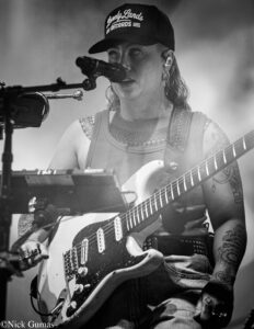 Female guitarist singing into a microphone while playing a white electric guitar on stage, tattoos visible on arms.