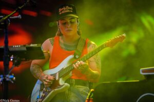Guitarist wearing an orange tank top and a cap reading 'Lonely Lands Records' plays a white electric guitar on a colorful stage.