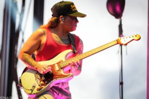 Guitarist wearing a red tank top and cap, playing a cream-colored electric guitar on stage.