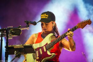 Female guitarist performing live on stage, wearing a red tank top and black cap, playing a mint-green electric guitar with a microphone and keyboard in view.