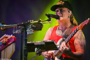 Musician in a red tank top wearing a cap, singing into a microphone while playing an electric guitar on stage with yellow-green lighting.