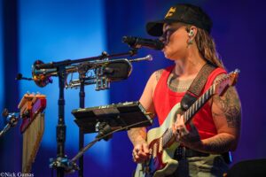 Musician in a red tank top playing an electric guitar on stage with blue lights in the background; visible tattoos on arms and neck.