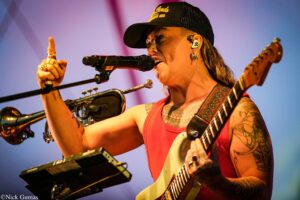 Musician on stage singing into a microphone while pointing with a raised finger and playing an electric guitar, with a purple backdrop.