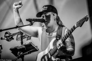 Black-and-white photo of a female musician performing on stage, singing into a microphone while playing an electric guitar, with her left arm raised.