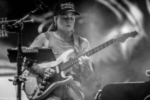 Female guitarist with tattoos playing an electric guitar on stage, wearing a cap and headset in a black-and-white photo.