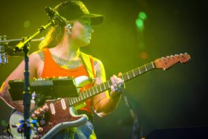 Guitarist in an orange tank top and cap playing a sunburst Fender Stratocaster onstage under yellow-green lights, tattoos visible on arms.