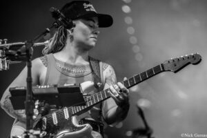 Female guitarist on stage wearing a cap, playing an electric guitar with a pedalboard nearby during a live show.