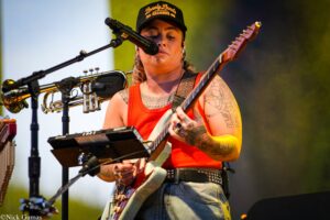 Musician with tattoos plays an electric guitar on stage, singing into a microphone, wearing a red tank top and a black cap.
