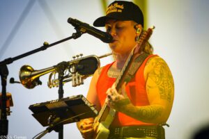 Musician in an orange tank top and cap sings into a microphone while playing an electric guitar, with tattooed arms visible on stage.