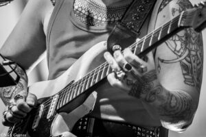Close-up of a tattooed guitarist's hands fretting and picking on an electric guitar in black and white.
