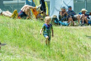 Young child running through tall grass toward the camera as adults sit and watch behind a chain-link fence in a sunny field.