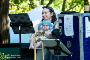 Woman holding a baby with blue headband and headphones in an outdoor control area, looking at a screen.