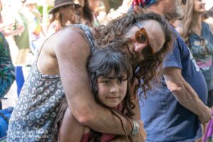A man with orange sunglasses embraces a young girl at an outdoor crowd event, smiling as they hug.