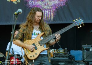Male bass guitarist with long hair and glasses, wearing a plaid shirt, playing a natural wood bass on stage with amps and gear around him