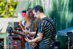 Guitarist in a tie-dye shirt playing an acoustic guitar at an outdoor event, with other band members softly blurred in the background.