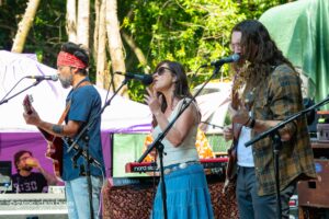 Outdoor band performance with a female lead singer and two guitarists on a sunny festival stage, microphones set up and keyboards in view