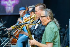 Saxophonist in a green shirt plays his instrument at a live band performance, with other brass players blurred in the background.