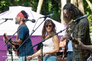 Three musicians perform on an outdoor stage with microphones and guitars; a woman in sunglasses stands center smiling.