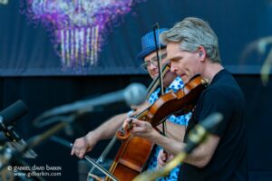 Two violinists perform on a stage, focusing on their bows and instruments, foregrounded by a man with gray hair and earplugs