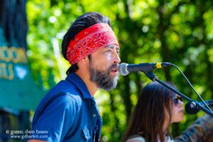 Man wearing a red paisley bandana and blue shirt singing into a microphone outdoors, with a green, blurred background.