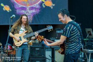 Two musicians performing on a stage: one with long hair and glasses playing a light wood bass guitar, the other smiling and playing an electric guitar, with amplifiers and gear around them during a live performance.