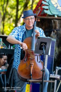 Male street musician playing a cello outdoors, wearing a blue hat and polka dot shirt, reading sheet music on a stand with a tablet nearby.