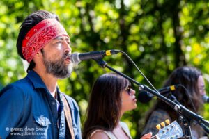 Man wearing a red bandana sings into a microphone during an outdoor acoustic performance, with green trees in the background.