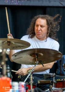 Drummer with long curly hair playing a drum kit, holding a drumstick above a cymbal during a performance