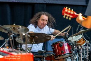 Drummer with long curly hair wearing a white shirt plays a red drum set on stage, cymbals nearby.