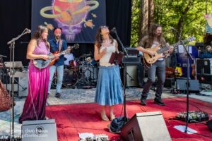 Outdoor band performance with a female vocalist center stage in a white top and blue skirt, flanked by guitarists and a drummer behind them.