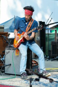 Guitarist in a blue shirt and red bandana playing a sunburst Gibson electric guitar on an outdoor stage with amps nearby.