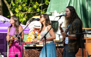 Three musicians perform outdoors: a woman in pink with a guitar, a center female singer at a microphone, and a man with long hair and a guitar, with tents in the background.