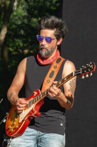 Guitarist with spiky gray beard wearing sunglasses and a red bandana, playing a red Les Paul-style guitar on an outdoor stage.