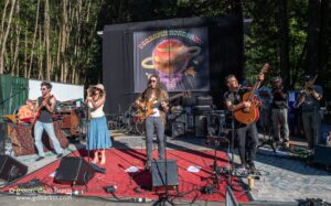 Outdoor band performance on a red carpet stage amid trees, with multiple musicians playing guitars, singing, and drums; colorful banner behind them.