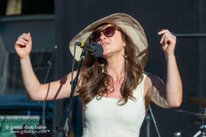 Female singer wearing a wide-brim straw hat and sunglasses, singing into a microphone on stage.