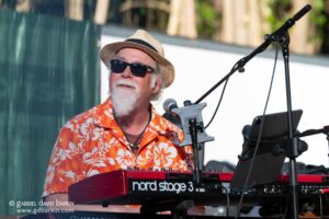 Musician wearing a straw hat and sunglasses playing a red Nord Stage 3 keyboard on stage outdoors, with microphone nearby and sheet music stand visible.