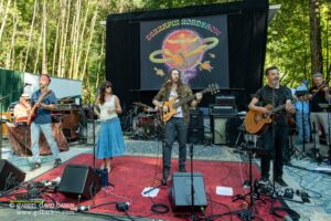 Outdoor band performs on stage in a park, with multiple musicians, guitars, drums, and microphones under a large banner.