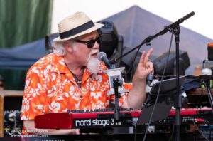 Musician with straw hat and sunglasses sings into a microphone while playing a red Nord Stage 3 keyboard on stage.