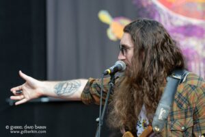 Musician with long hair and sunglasses singing into a microphone on stage, wearing a plaid shirt and guitar strap, arm extended toward the audience.