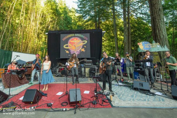 Outdoor forest concert with a full band on a stage, singer at center, multiple instrumentalists, and a Paper Moon Presents banner above.