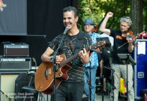 Male guitarist sings into a microphone while playing an acoustic guitar on an outdoor stage, with bandmates and trees in the background.