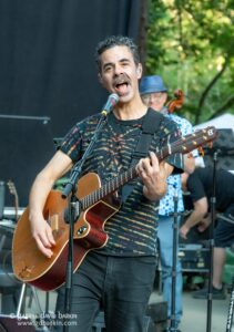 Male musician singing into a microphone while playing a sunburst acoustic guitar on an outdoor stage, wearing a tie-dye shirt and a strap across his chest.