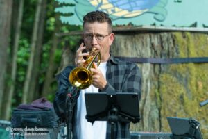 Man playing a brass trumpet outdoors, wearing glasses and a plaid shirt, with a music stand in front.