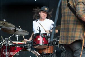 Drummer with long curly hair in a white T-shirt and black cap playing a red drum kit on stage, drumsticks in hand.