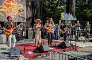Outdoor concert with a multi-member band performing on a stage; guitarist and vocalist front center, brass players in the background, Paper Moon Presents sign in trees nearby.