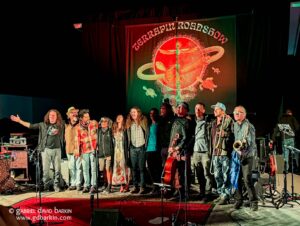 A diverse group of musicians and singers on a stage, holding instruments and singing together, with a large red-and-green banner behind them.