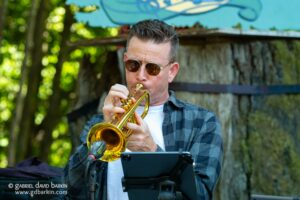 Male musician playing a trumpet outdoors, wearing sunglasses and a plaid shirt, focused on his instrument near a music stand.
