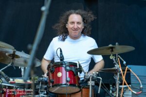 Drummer with long curly hair in a white shirt playing a red drum set on stage.