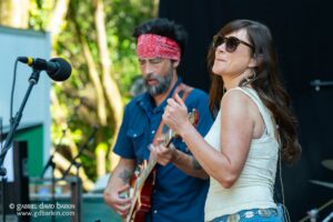 Female singer wearing sunglasses and a white lace top plays guitar on an outdoor stage, with a bearded man in a red bandana accompanying in the background.